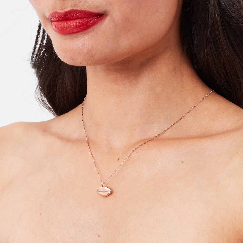 Close-up of a woman's chest and neck, showcasing a rose gold necklace with a lip-shaped pendant against a white background.