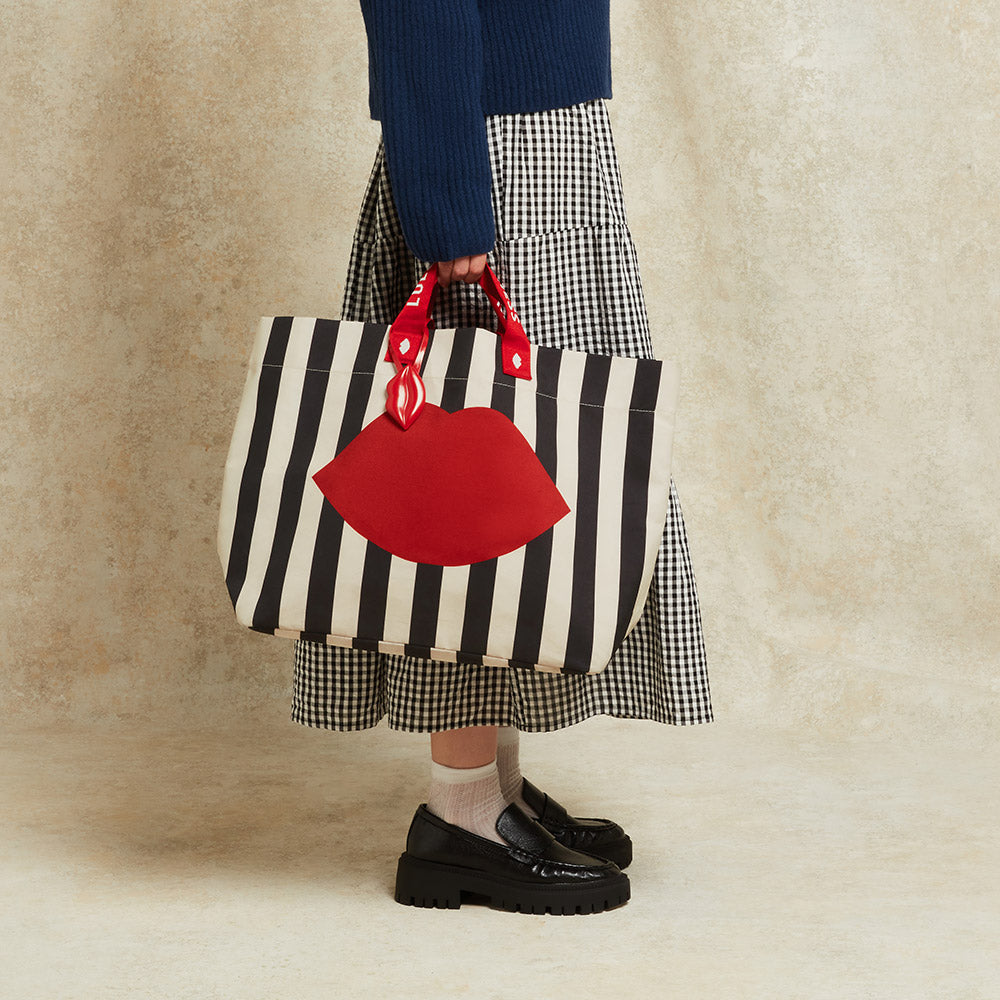 Woman holding a black and beige striped bag with red lips on the front against a beige background.