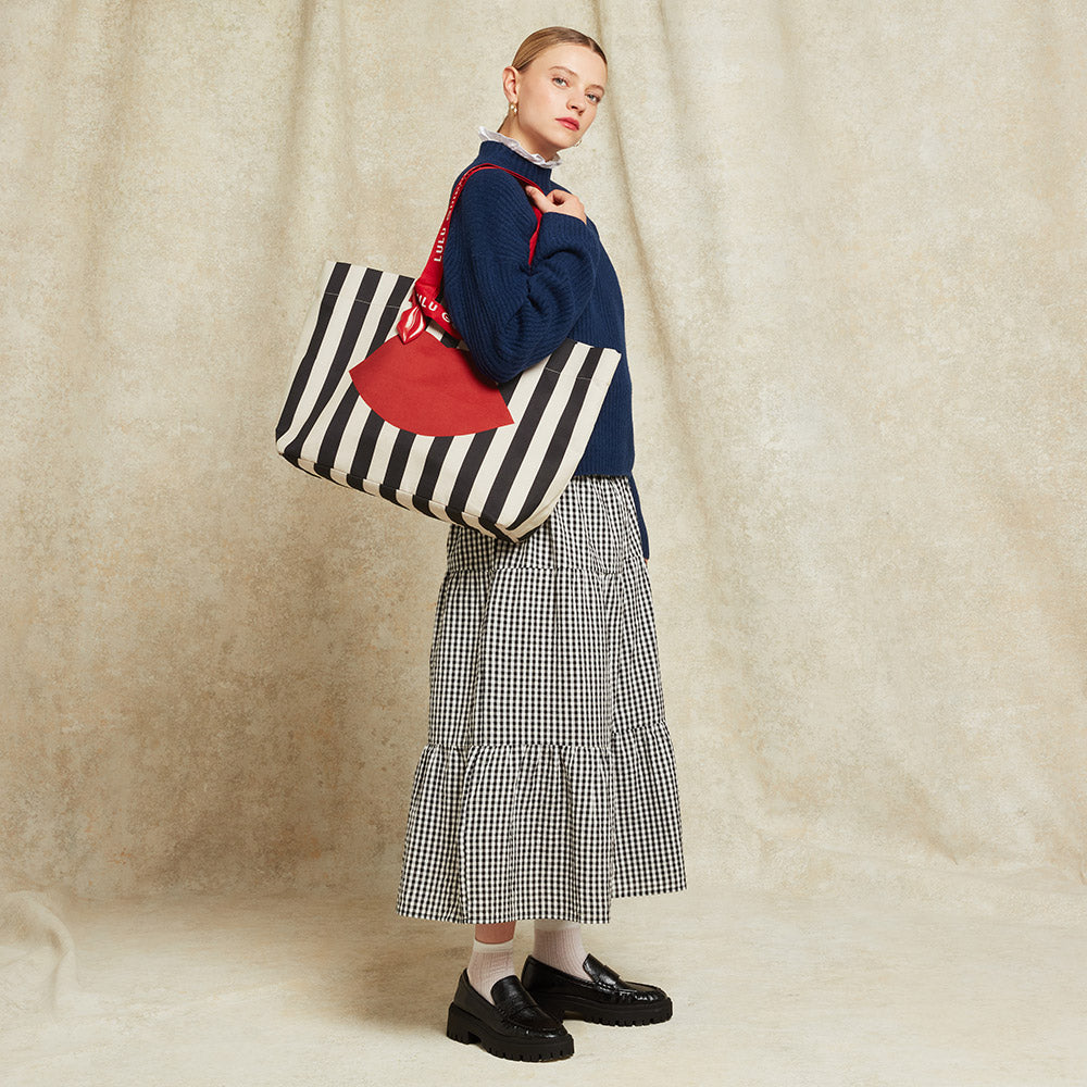 Woman holding a black and beige striped tote bag with a red lip printed on the front against a beige background.