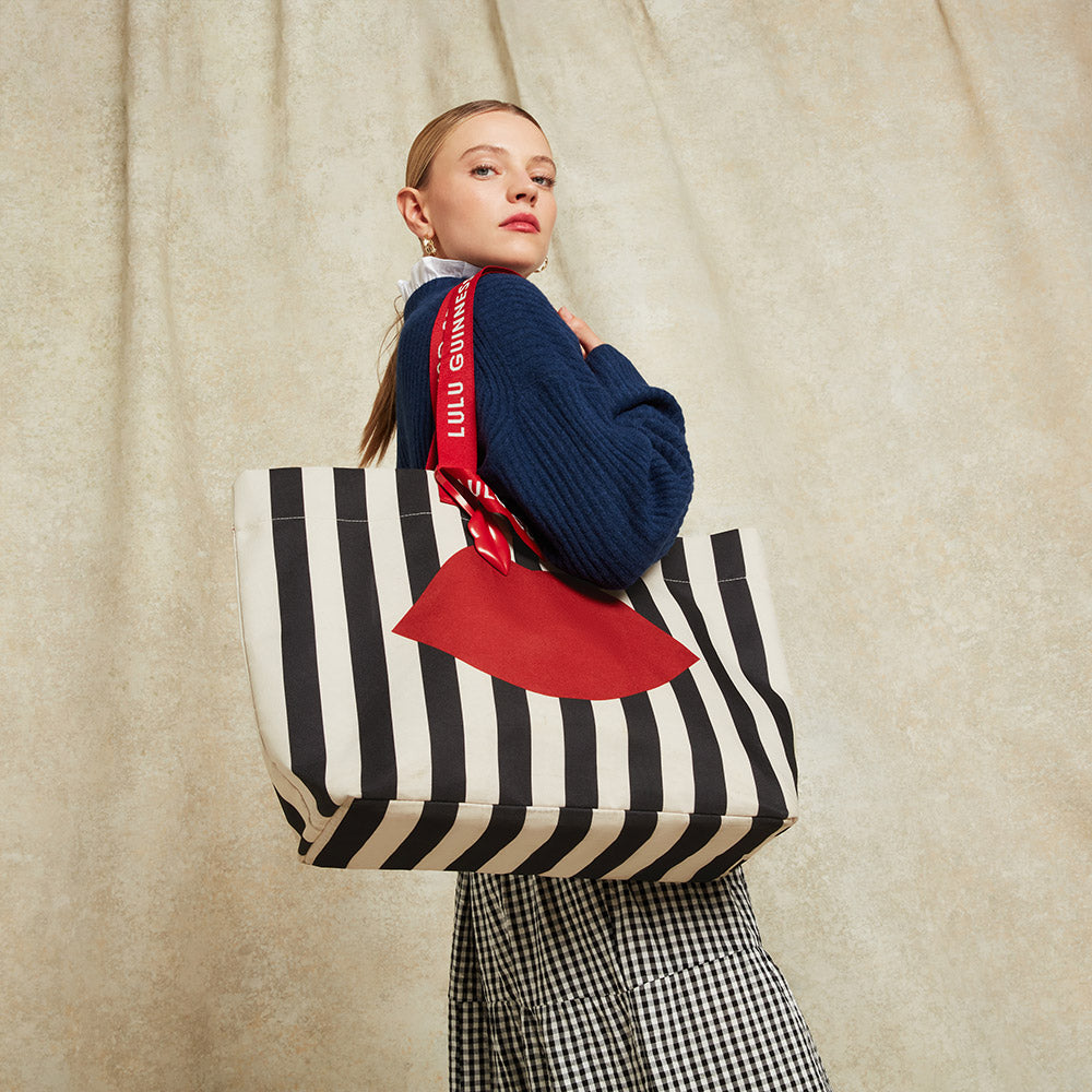 Woman holding a black and beige striped tote bag with a red lip printed on the front against a beige background.