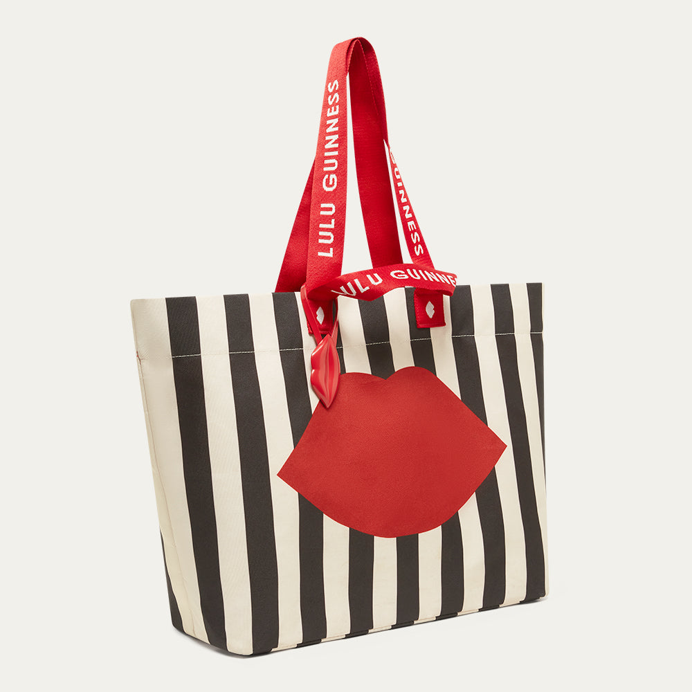 Striped black and beige tote bag with red lip accents and 'Lulu Guinness' branding on the strap on a white background