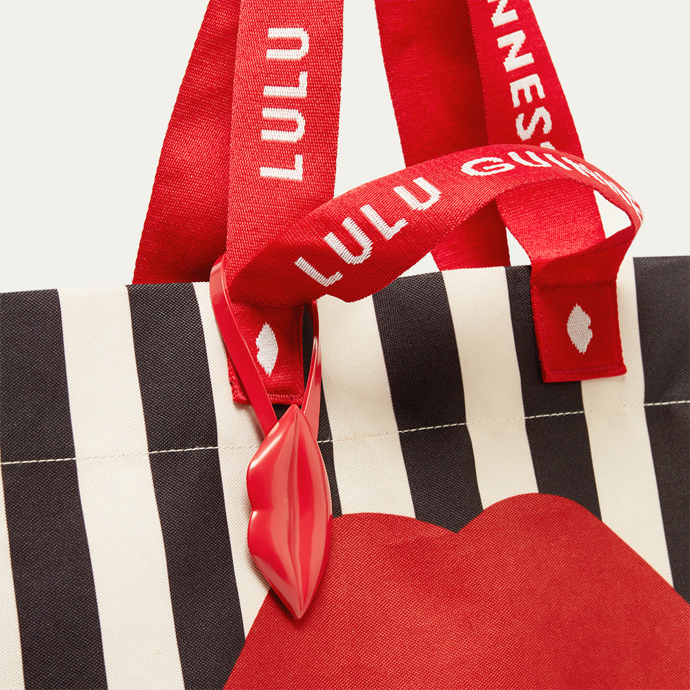 Close up image of a black and beige stripe bag with red lip detailing and 'Lulu Guinness' branding on the strap.
