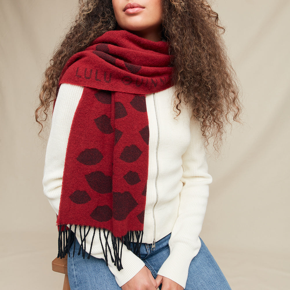 Person wearing a red scarf with black lip pattern and 'Lulu Guinness' branding, standing against a beige background.