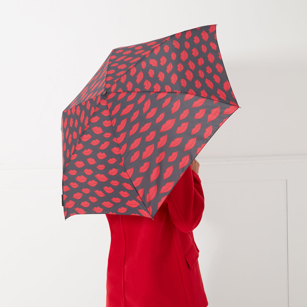 A woman in a red coat holds a dark gray umbrella with a pattern of red lips printed on it.
