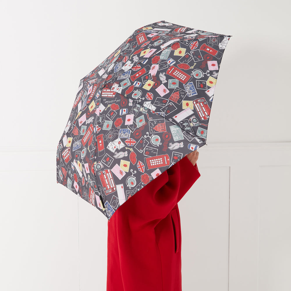 Compact umbrella with a dark background and a colorful pattern of postage stamps, red lips, London landmarks and other related images. A person wearing a red coat holds the umbrella.
