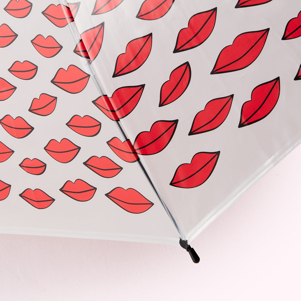 Close-up of a clear umbrella with a pattern of red lips printed on a light gray background.
