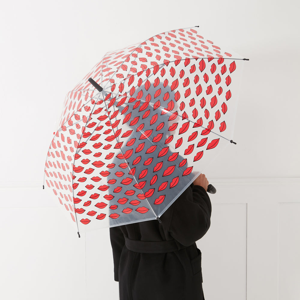 Person from behind holding a clear umbrella with a red lips print against a white background.
