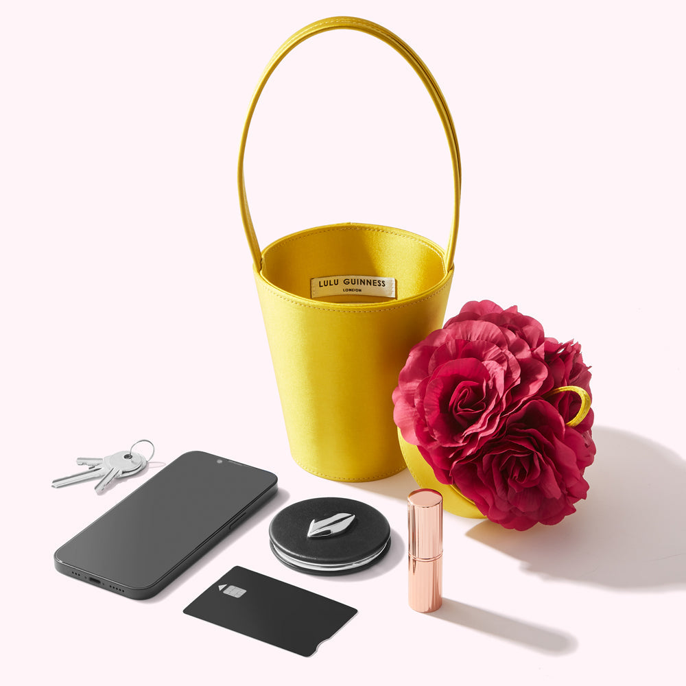 A yellow Lulu Guinness bucket bag with a fuchsia rose detail sits next to a smartphone, keys, a credit card, and lipstick on a white background.