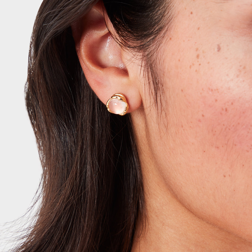 Close-up of a person's ear wearing gold stud earrings with pale pink stones.
