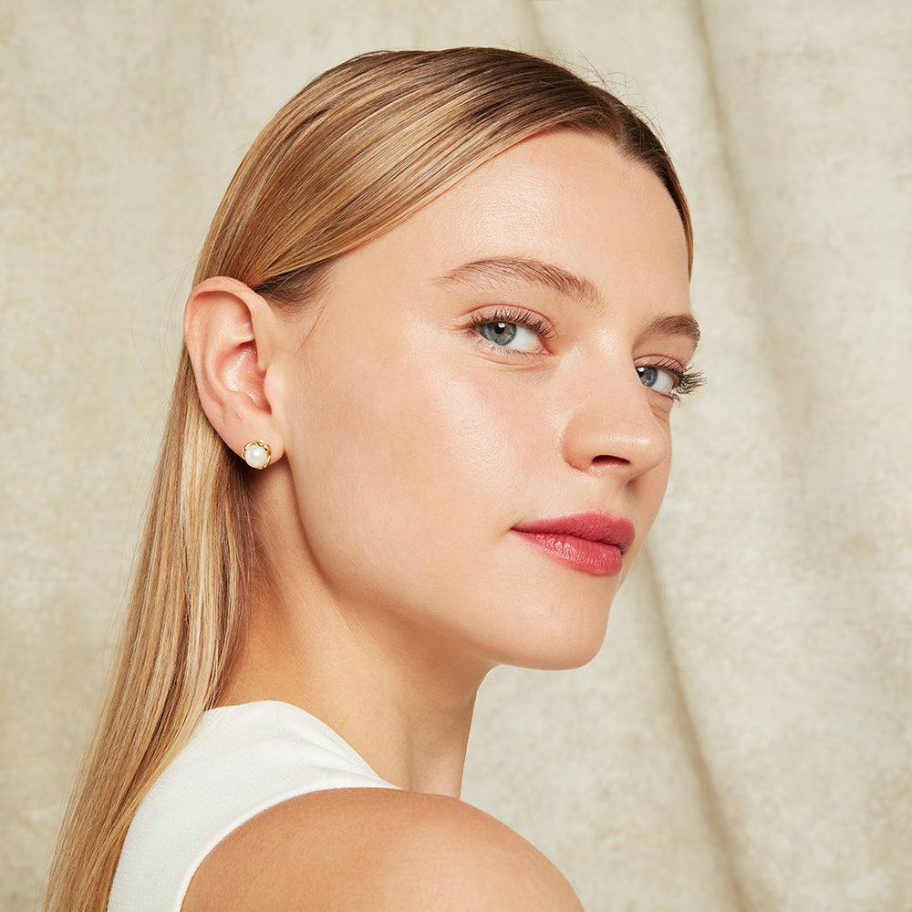 Close-up of a woman wearing gold and pearl stud earrings with a neutral background