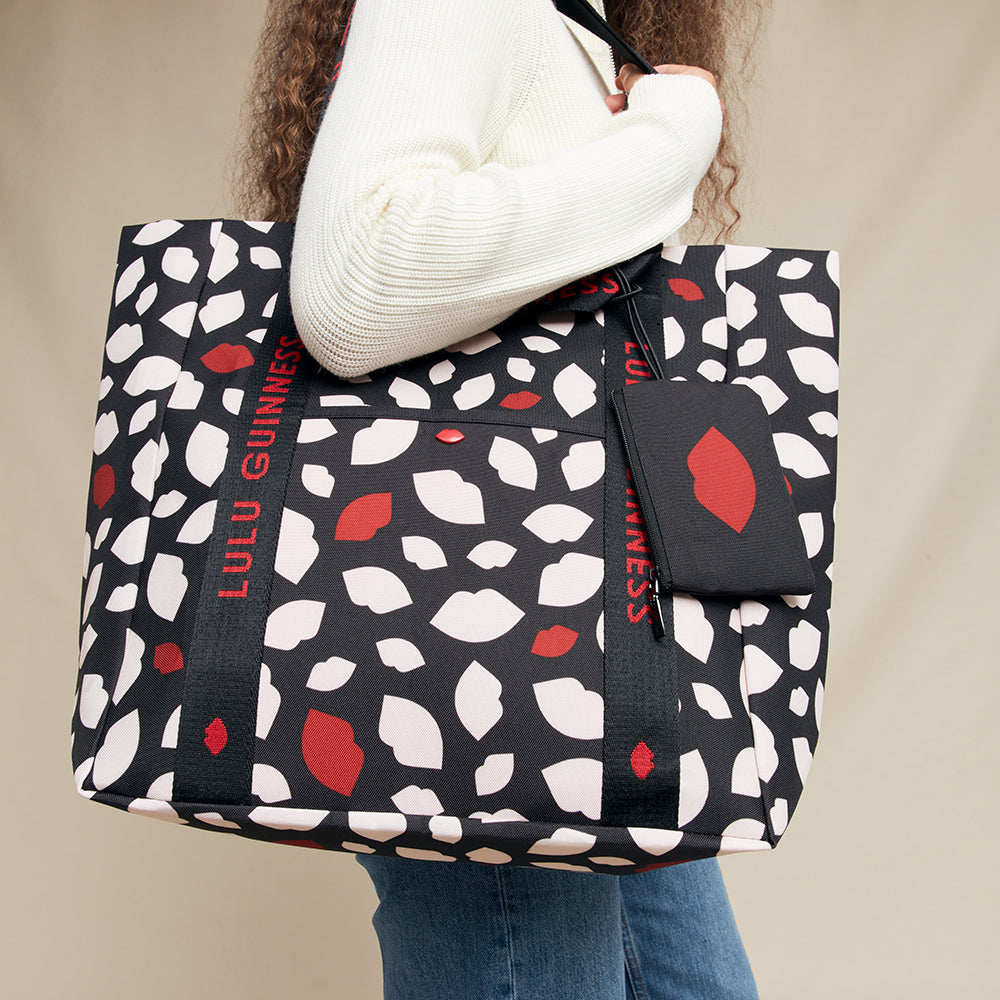 Person holding a black tote bag with red and white lip pattern, featuring 'Lulu Guinness' branding.
