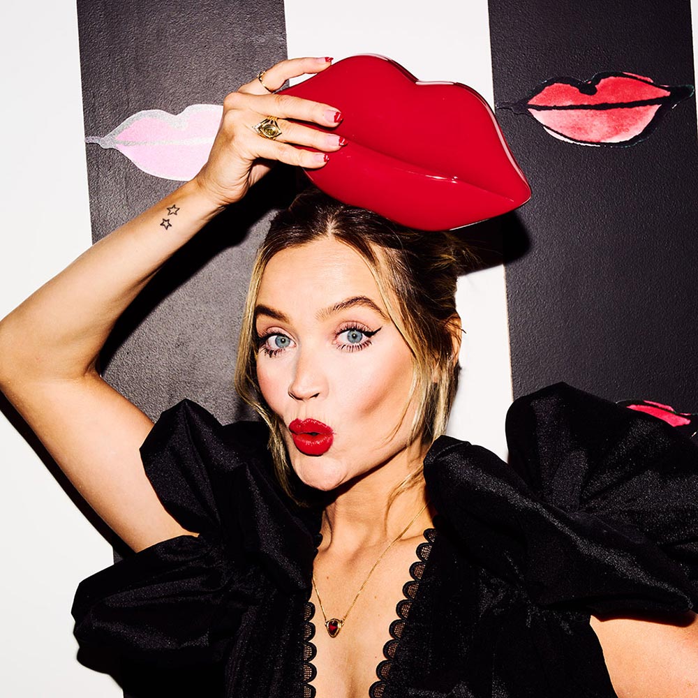 Laura Whitmore in red lipstick and wearing a black ruffle top holding a red lips clutch above her head.