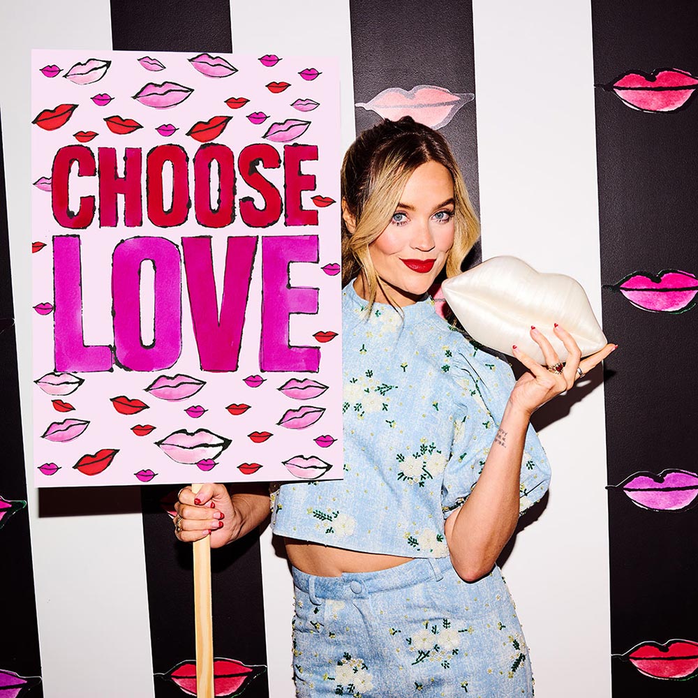 Laura Whitmore in a blue floral outfit holding a pearl lips clutch and standing next to a Choose Love with Lulu Guinness sign.