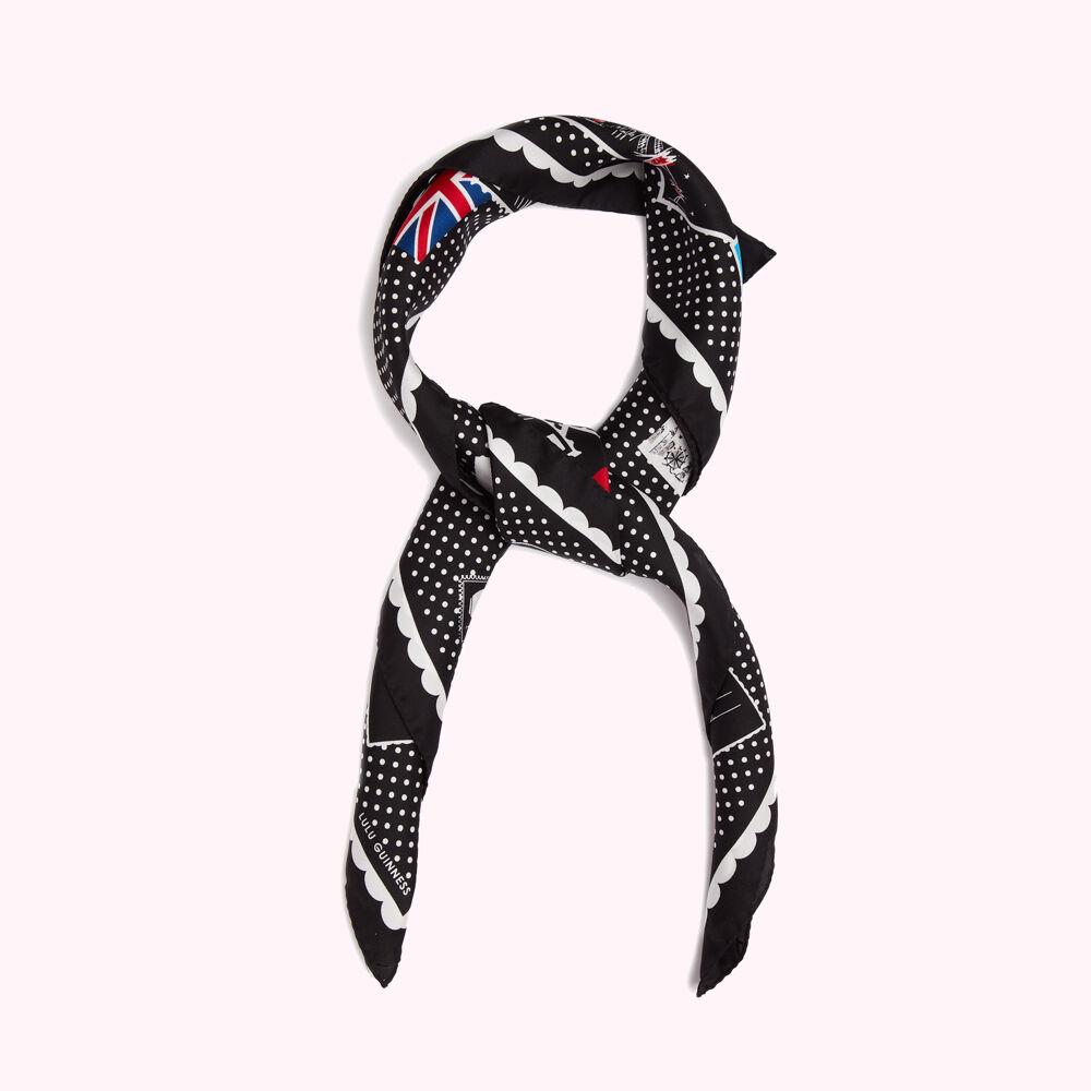 Black silk scarf with white polka dots and a repeating postage stamp print featuring the Union Jack and other iconic imagery, tied in a knot. The brand name "Lulu Guinness" is visible on the scarf.
