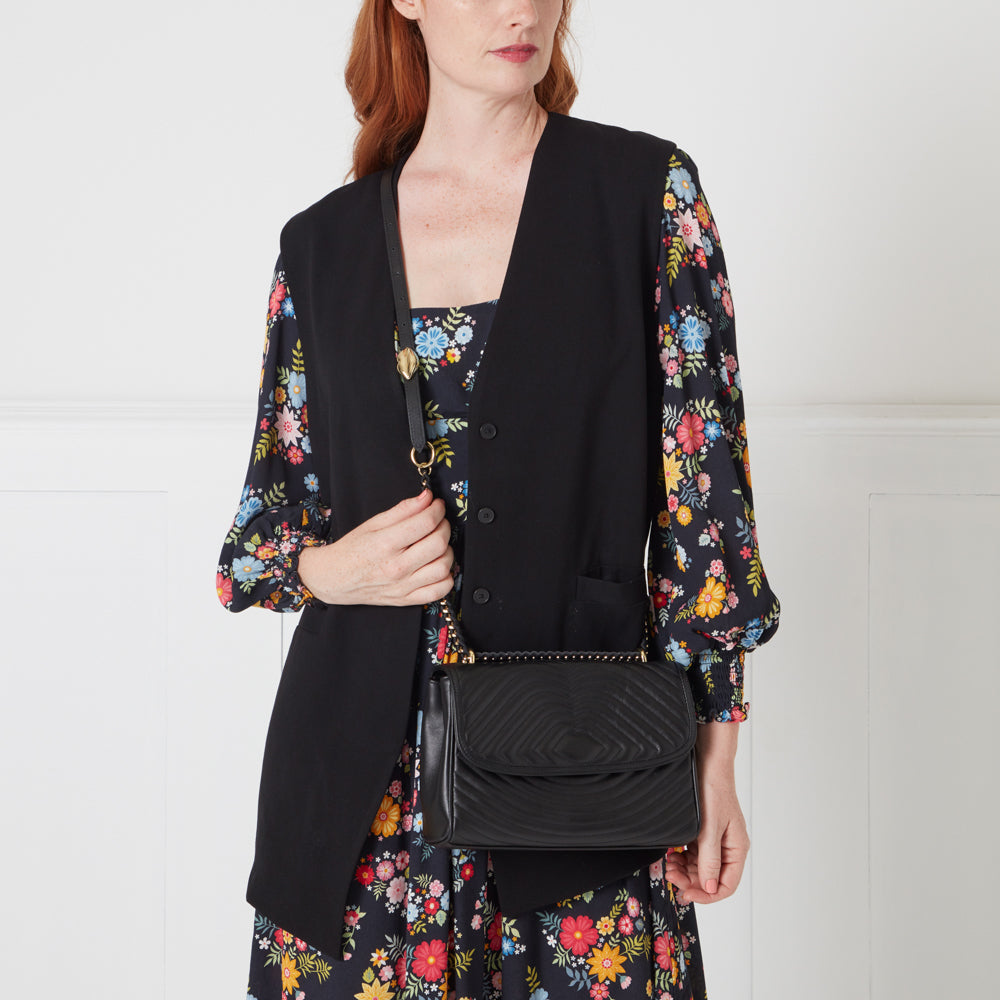 Woman wearing a black vest and floral dress holding a black quilted crossbody bag.
