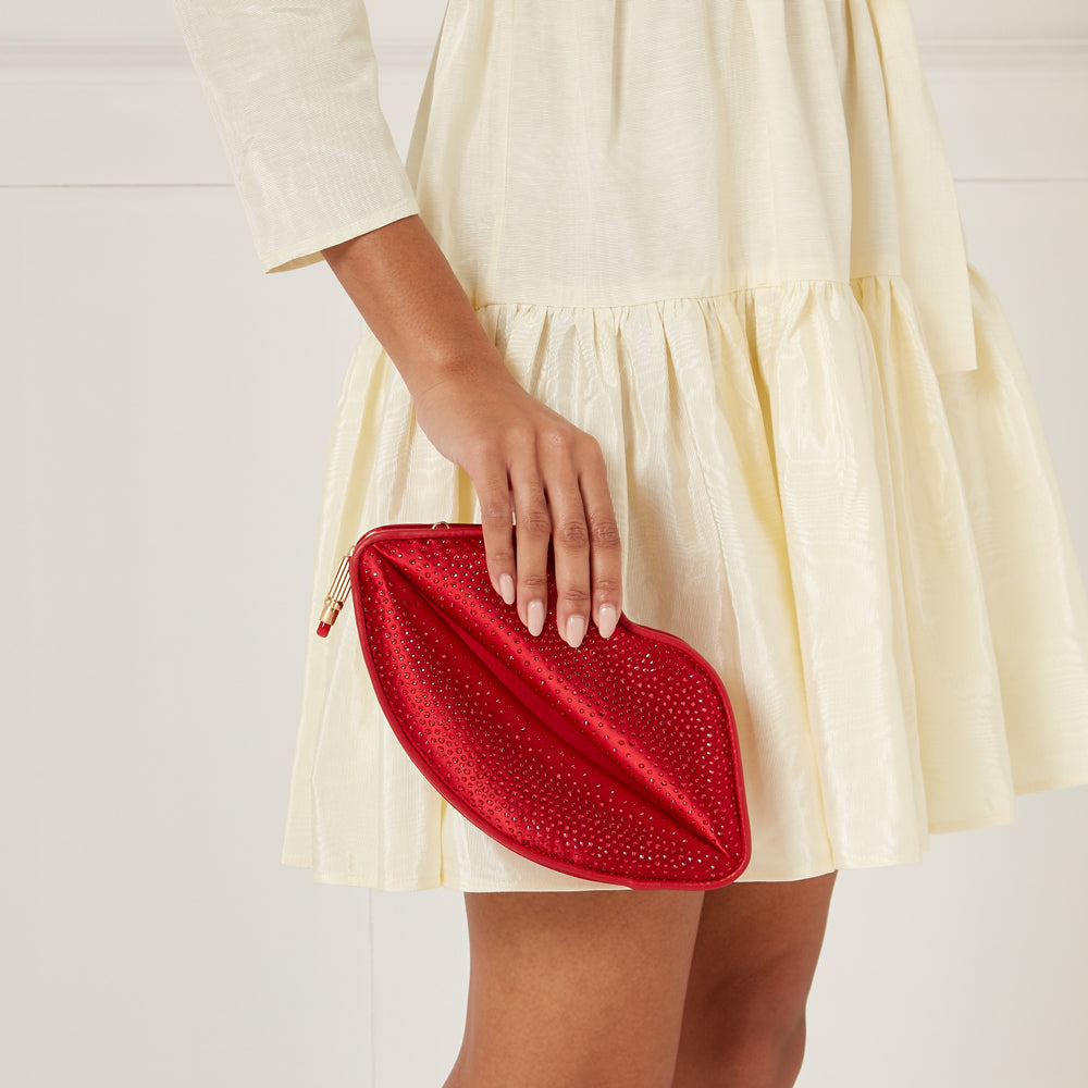 A woman in a pale yellow dress holds a red, lip-shaped clutch encrusted with rhinestones.